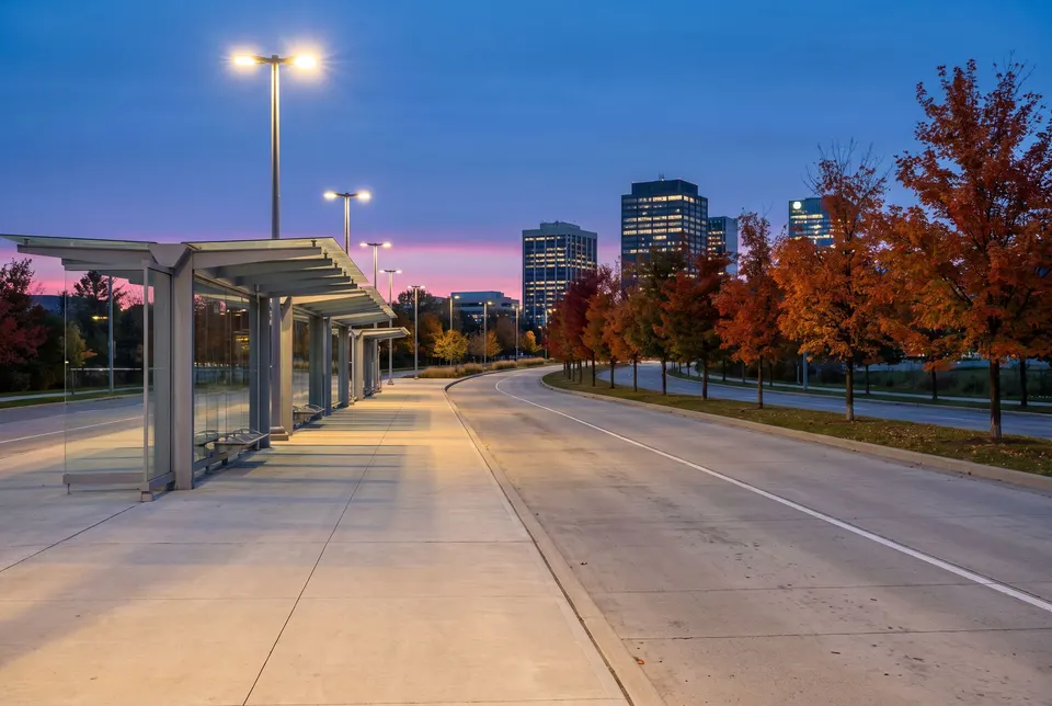 Une station d’autobus moderne et vide au crépuscule : quai de béton, abris de verre, voie réservée qui tourne, érables orangés et tours éclairées au loin.